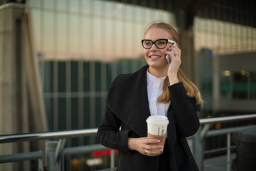 Happy smiling woman skilled manager talking via mobile phone while standing with take away coffee outside company during break at job. Joyful female with good mood having cell telephone conversation