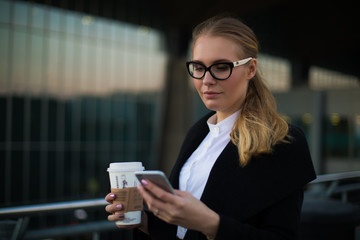 Female smart university student in glasses reading text message on mobile phone while standing with take away coffee outdoors. Woman secretary received notifications on cell telephone gadget