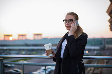 Woman proud executive director in glasses and coat having cell telephone conversation with partner while standing outside company in evening time. Serious female manager talking via mobile phone