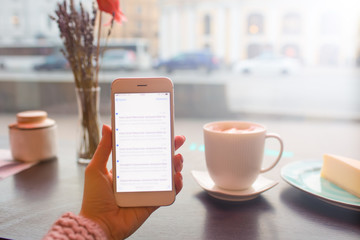 Closely female blogger holding mobile phone with blurred notifications on the screen while sitting in coffee shop with cup of latte during recreation time in holidays