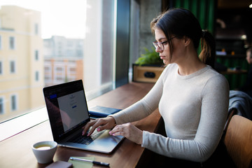 Serious woman in stylish casual wear and spectacles professional business content writer searching information in internet via pc laptop computer, sitting in coffee shop. Hipster girl using apps