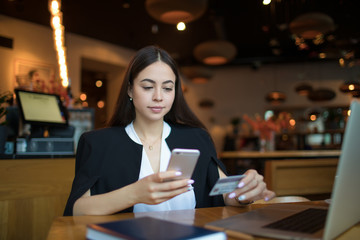 Proud woman government worker in formal wear online payment via cell telephone using credit debit card while sitting with netbook in luxury restaurant. Female entrepreneur booking via mobile phone