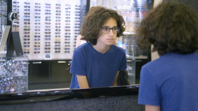 choice of frames for glasses in optics. Mom and her teenager son choose a frame for glasses while looking in a large mirror in optics.