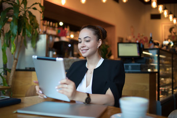 Happy smiling woman hostess reading good new on website via portable touch pad while sitting in modern restaurant during work break. Joyful female online shopping store via digital gadget