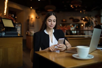 Young prosperous business woman online searching information on account via mobile phone while sitting in luxury restaurant during work break. Female jurist using cell telephone for chat in internet