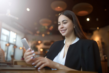 Beautiful smiling business woman sending sms via mobile phone while sitting in modern restaurant during recreation time. Happy joyful female blogger online shopping store via cellphone during resting