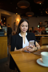 Charming woman smart college student watching photo on mobile phone during recreation time in restaurant. Confident female with good mood connection to internet via cell telephone, sitting in cafe
