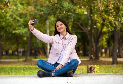 Brunette girl photographs herself on a cell phone while sitting in an autumn park.