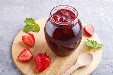 Strawberry jam in a glass jar with berries and leaves on gray concrete background, side view.