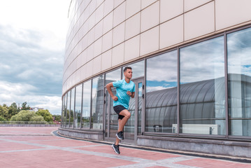 A man runs in the summer in the city for a fitness training in sportswear, a T-shirt and sneakers, free space for text. Glass building window background. Active lifestyle of youth.