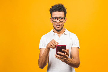 Smiling black young man in glasses holding phone isolated on yellow studio background with copy space aside, african guy using mobile applications, texting.