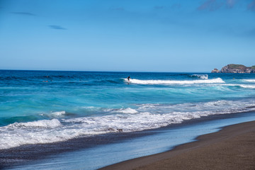 Fototapeta na wymiar Beautiful landscape surfers riding a wave on Santa Barbara Beach, Black Sand Beach, Ribeira Grande, Sao Miguel Island, Azores, Portugal