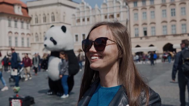 Beautiful girl in sunglasses stands on the town square and photographs historic buildings. A man in a panda costume in the background