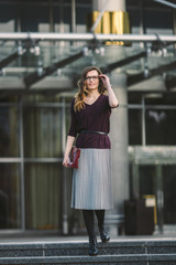 Fototapeta premium Business woman standing on street against office building. City business woman working. Portrait business woman smiling with glasses. Woman posing outside office building summer business outfit