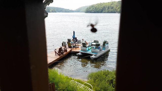 A group of people sunbathing on a wooden pier, with a young man jumping into the water seen from a wooden valcon with a small spider working on his cobweb