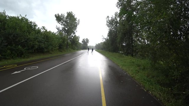 Man and a woman are rollerblading during the rain. View from the back, removed. Park, summer.