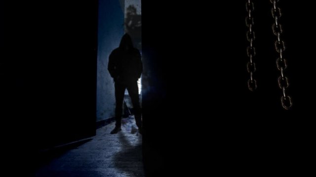 Hooded Man Behind Opening Door 4K Loop features a dark room with hanging chains swinging and a door opening to reveal a man in a black hooded and then closing again in a loop