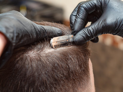 Hair cutting with open razor, close up view. Hands in black rubber gloves with straight razor. Hairdressing stylist at work in barbershop. Selective soft focus. Blurred background
