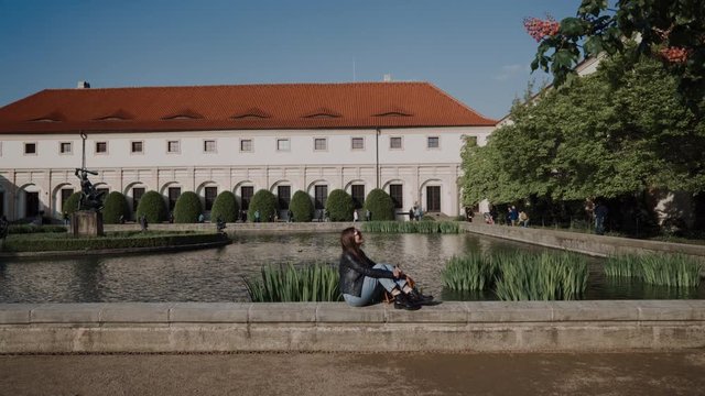 Pretty young lady with backpack sitting on parapet of pond. There are Wallenstein Palace and the pond with fish and sculptures on background, currently the home of the Czech Senate in Prague, Czech
