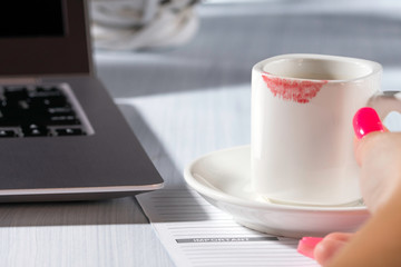 Cup of coffee and laptop, lipstick mark on cup, office desk, woman hand