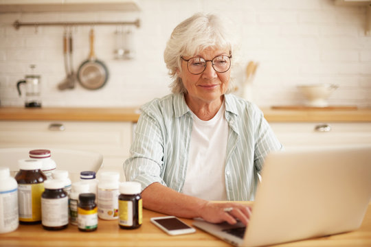 Indoor Shot Of Happy Positive Senior Woman With Gray Hair Sitting At Kitchen Table, Surrounded With Bottles Of Vitamins, Ordering More Online Using Portable Computer. Food, Nutrition And Diet