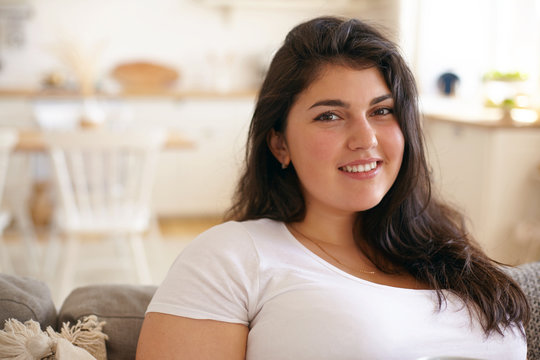 Close up indoor picture of attractive cute young female with large curvy body, chubby and broad shoulders posing indoors, relaxing on couch in living room, looking at camera with joyful smile