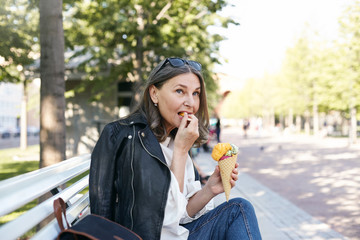 Food, travel, people and lifestyle concept. Side view of confident stylish middle aged female enjoying happy peaceful days of her retirement, sitting comfortably on bench, eating ice cream