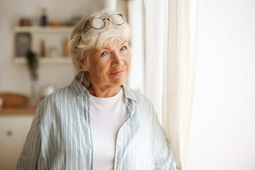 Aged people, maturity, retirement and lifestyle concept. Indoor image of casually dressed elderly mature female with gray hair standing by window, wearing glasses on her head, feeling lonely