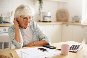 People, age, technology and finances. Depressed unhappy retired woman paying domestic bills online, trying hard to make both ends meet, sitting at kitchen table, surrounded with papers, using gadgets - obrazy, fototapety, plakaty