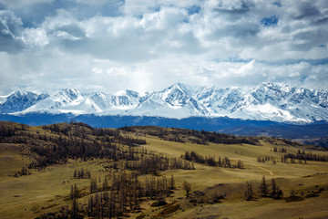 Fototapeta premium Fantastic mountain landscape. Rocky mountains with snowy peaks, hills covered with grass in the Alpine scene on a bright autumn day with blue sky and clouds. View of steppe and snow-covered mountains.