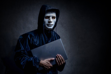 Anonymous hacker in white mask holding a laptop in the dark. Data thief, cyber crime concept....