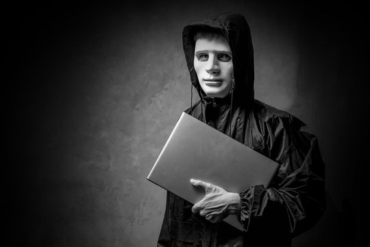 Anonymous hacker in white mask holding a laptop in the dark. Data thief, cyber crime concept. darknet and cyber security concept.