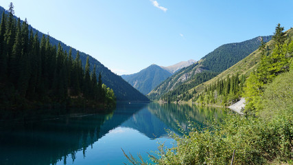 Kolsay lake located in the mountains of Kazakhstan. Turquoise water as a mirror. The water reflects the mountains, green hills, tall spruce, grass, sky and clouds. Beautiful landscape of mountain lake