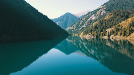 Fototapeta premium Kolsay lake located in the mountains of Kazakhstan. Turquoise water as a mirror. The water reflects the mountains, green hills, tall spruce, grass, sky and clouds. Beautiful landscape of mountain lake