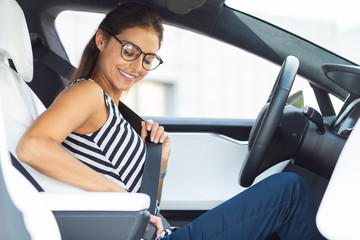 Woman wearing glasses sitting in her car and fastening seat belt