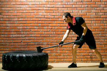 against a brick wall, a man in a black sports uniform, is engaged in cross-fit with a rubber tire and a hammer, there is a place for inscription