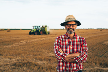 Naklejka na ściany i meble portrait of middle aged rancher standing crossed arms outdoors, agricultural machine in blurred background