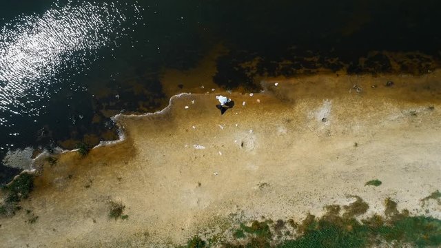 Aerial view of a man in a protective suit and a respirator who collects plastic trash on the banks of a dry and polluted river. Ecological catastrophy. Anthropogenic influence. 4K drone footage.