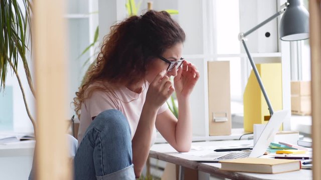 Medium shot of Caucasian businesswoman with long curly hair sitting at desktop at laptop, putting on eyeglasses, looking at laptop and making note in notebook