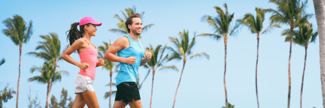Healthy people fit active lifestyle couple running on palm trees tropical background panoramic banner. Happy friends exercising together -training buddy friend.