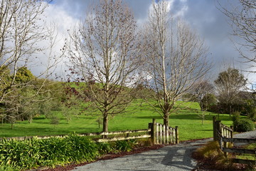Gravel road leading to countryside property through open wooden gate with trees on side.