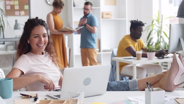 Pan shot of Caucasian businesswoman in casual clothes sitting at laptop with her legs on desk, having online video conversation while her multiethnic coworkers working on background