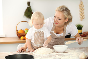 Little girl and her blonde mom in beige aprons playing and laughing while kneading the dough in kitchen. Homemade pastry for bread, pizza or bake cookies. Family fun and cooking concept