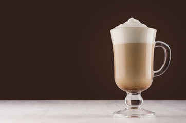 Latte coffee in elegant transparent glass with foam in modern dark brown kitchen interior, copy space.