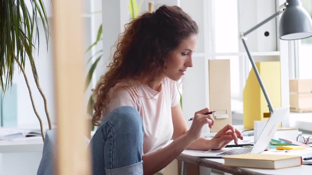 Medium shot of Caucasian businesswoman with long curly hair sitting at desktop, typing on laptop and making notes in notebook