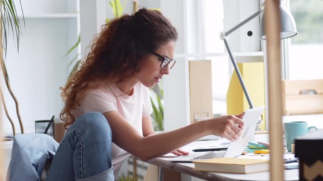 Medium shot of Caucasian businesswoman with long curly hair sitting at desktop at laptop, putting on eyeglasses, looking at laptop and making note in notebook