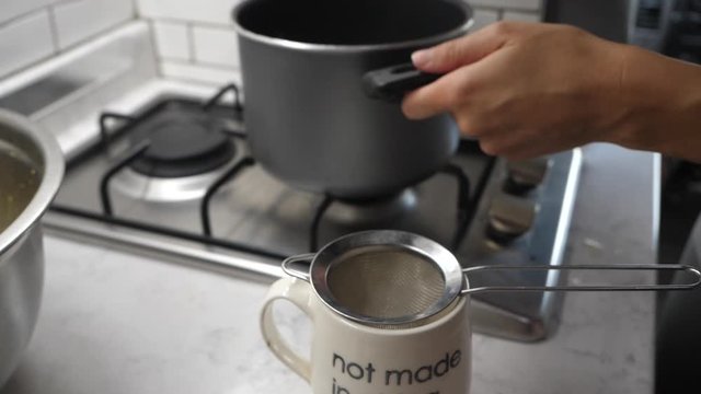 Person removes pot of soup from stove and filters broth into a mug.
