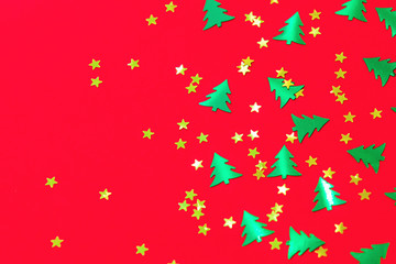 Green metallic foil christmas trees and gold stars confetti sparse on red background. Simple holiday concept. Design template. Frame with copy space. Winter festive backdrop. Top view, flat lay.
