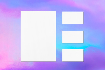 One empty white vertical a4 sized poster and three horizontal rectangle business card mockups with soft shadows on holographic background. Flat lay, top view