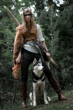 Art photo of a girl hunter with bow and husky on a background of dark forest